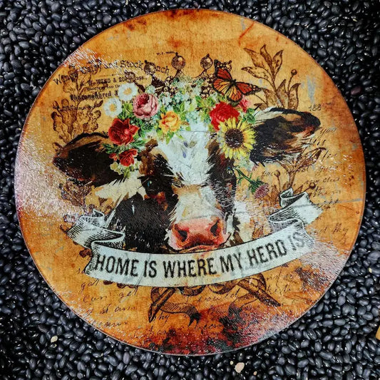 Round decorative glass cutting board with a cow illustration, flowers, and text on a textured surface. - unique housewarming gift
