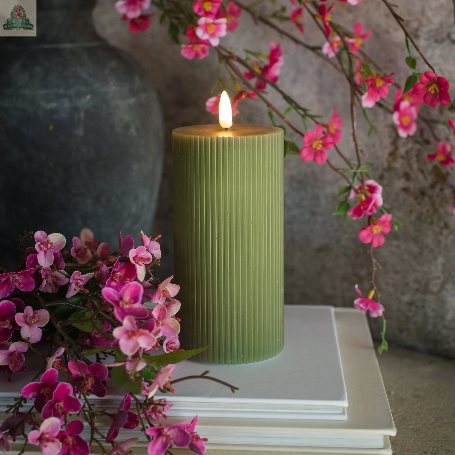 A 6" DARK GREEN GROOVED 3D FLAME CANDLE glows atop stacked white books, surrounded by vibrant pink flowers, with a gray vase and textured wall in the background.
