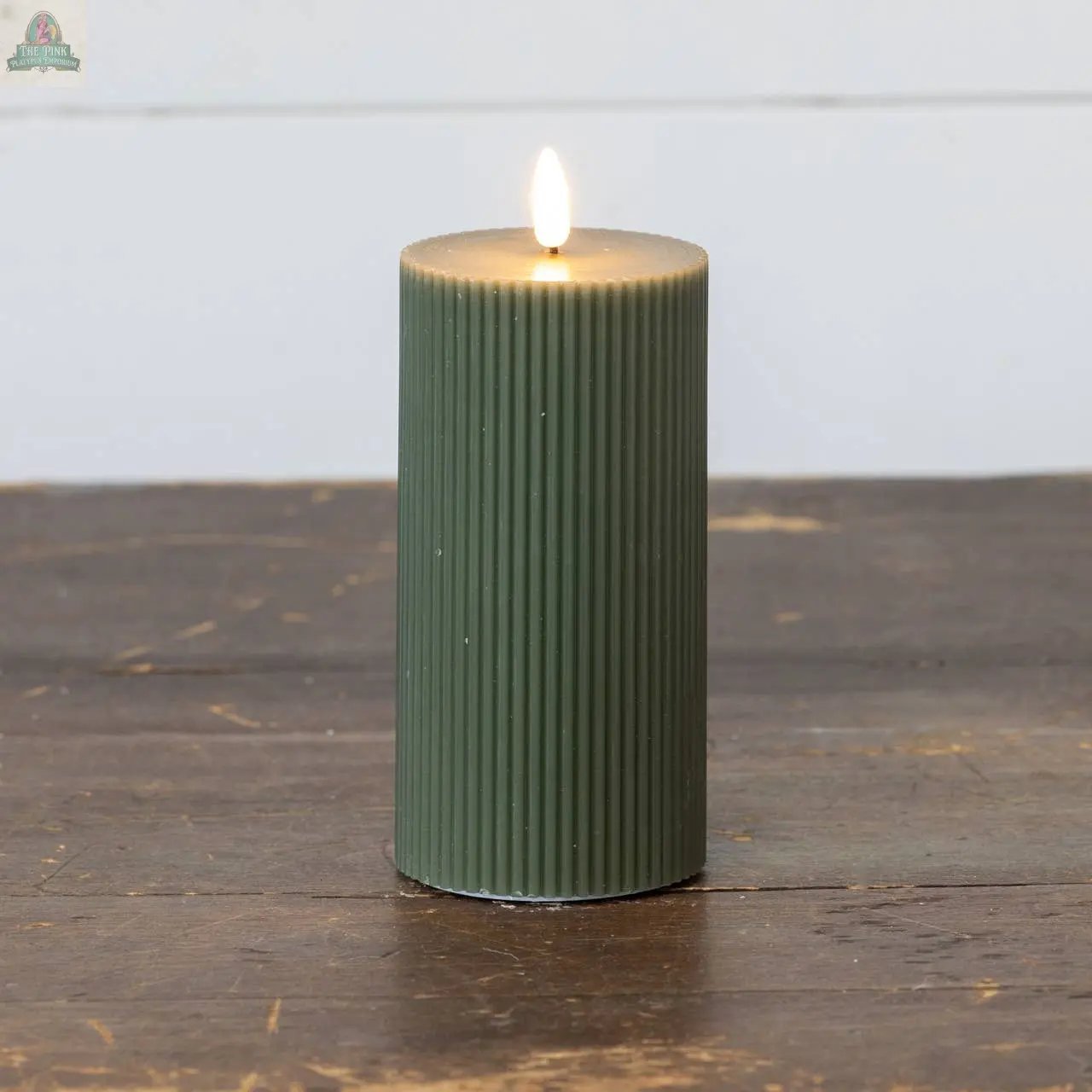 A 6" DARK GREEN GROOVED 3D FLAME CANDLE by Ragon House glows on a rustic wooden surface, highlighting its lifelike flameless design against a light background.
