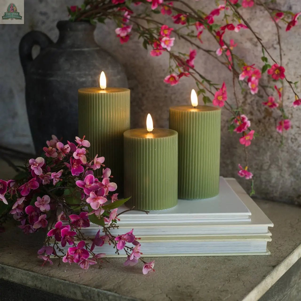 Three 6" DARK GREEN GROOVED 3D FLAME CANDLES are lit and arranged on stacked white books with pink flowers, while a dark vase with pink flowering branches sits in the background against a gray textured wall.