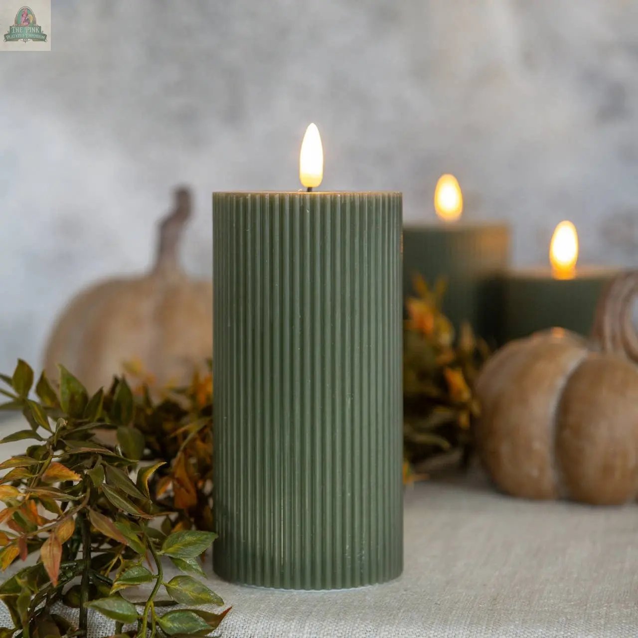 A 6" Dark Green Grooved 3D Flame Candle from Ragon House is lit among autumn leaves and pumpkins on a light surface, while two more flameless candles of the same style glow softly in the background.