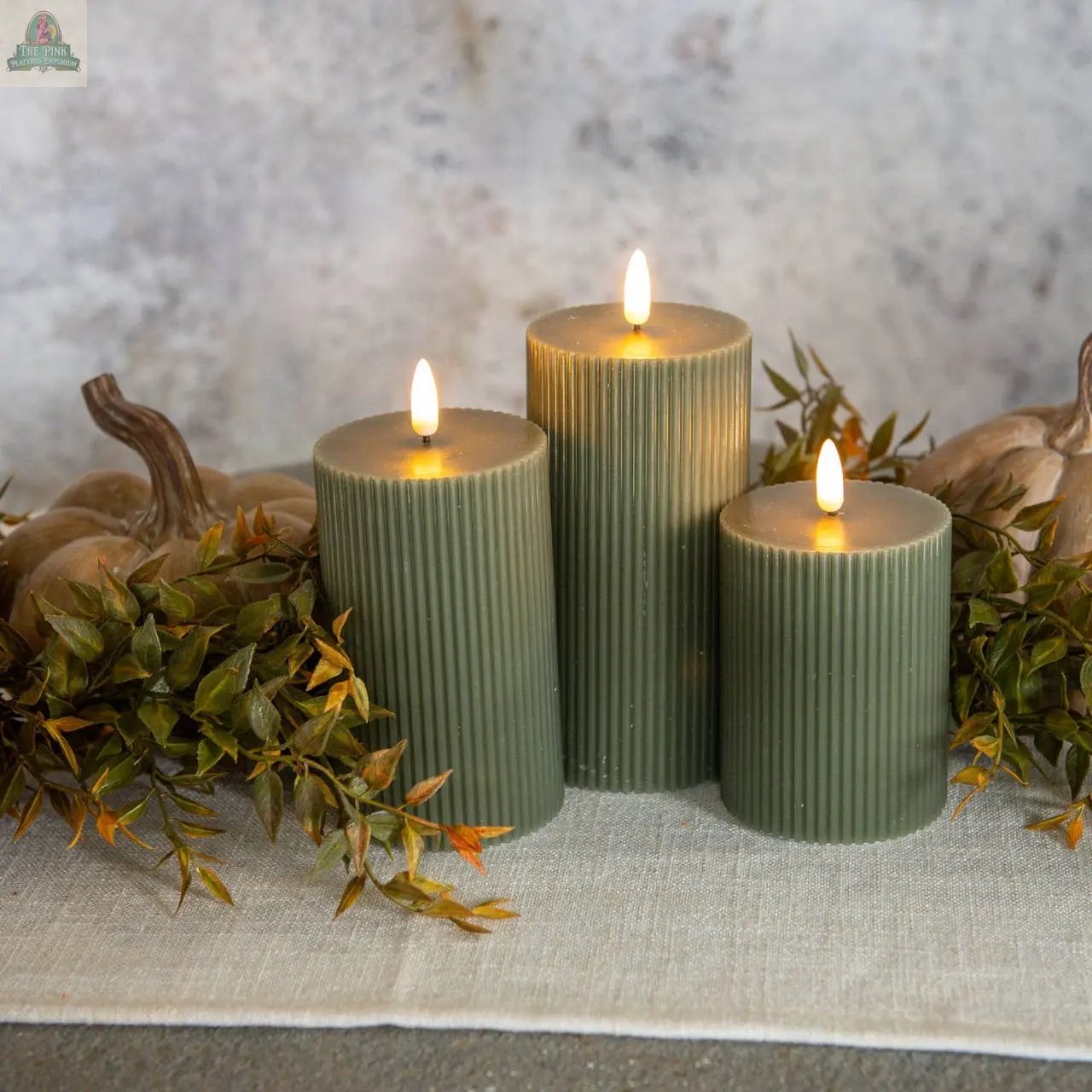 Three 6" Dark Green Grooved 3D Flame Candles are lit and arranged on a light runner with artificial leaves and decorative pumpkins, set against a neutral background.