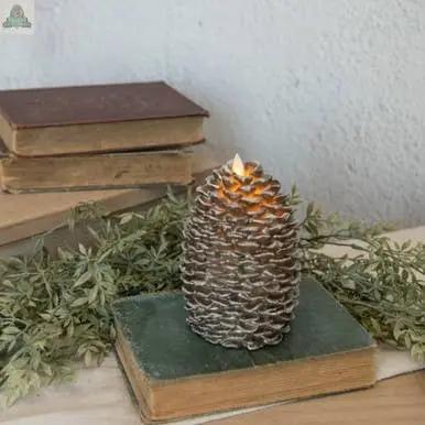 A 6" MOVING FLAME PINECONE CANDLE rests on a closed green book amid leafy branches, with two stacked books behind it—a charming piece of home decor set against a light-colored wall.