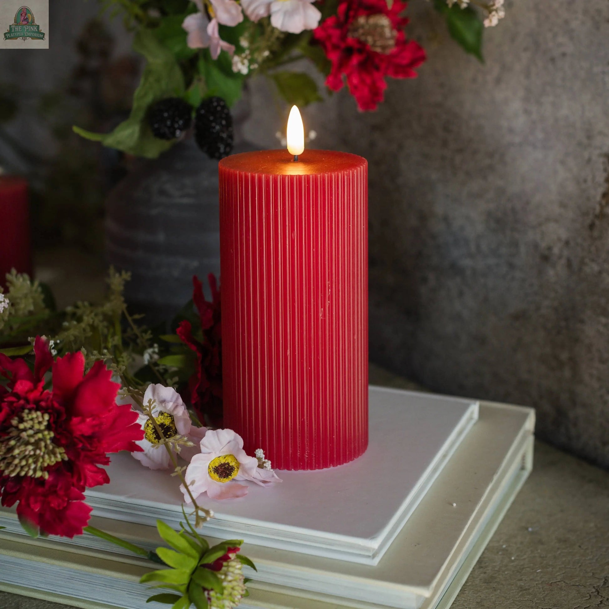 A 6" RED GROOVED 3D FLAME CANDLE with a lifelike, flameless glow sits atop two white books, surrounded by red and white flowers and greenery, against a gray background.