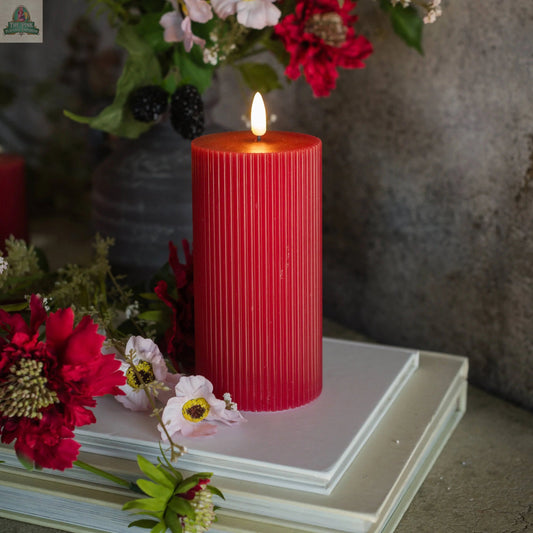 A 6" RED GROOVED 3D FLAME CANDLE with a lifelike, flameless glow sits atop two white books, surrounded by red and white flowers and greenery, against a gray background.