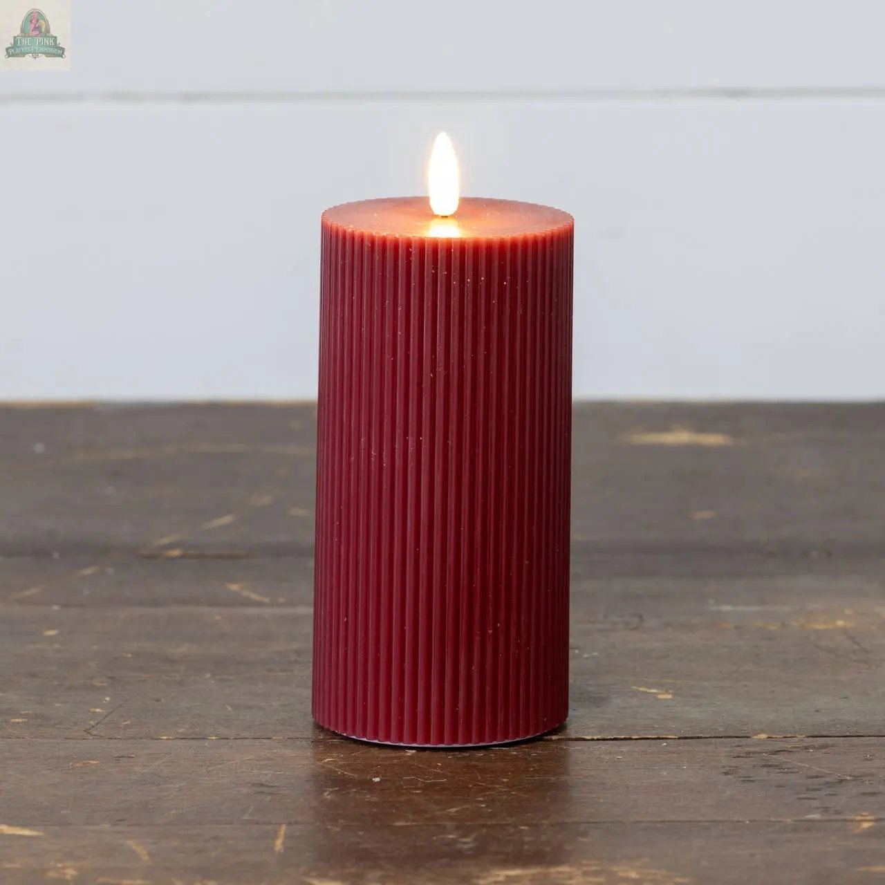 A 6" RED GROOVED 3D FLAME CANDLE sits lit on a dark wooden surface, its ribbed red design glowing warmly against the white wall.