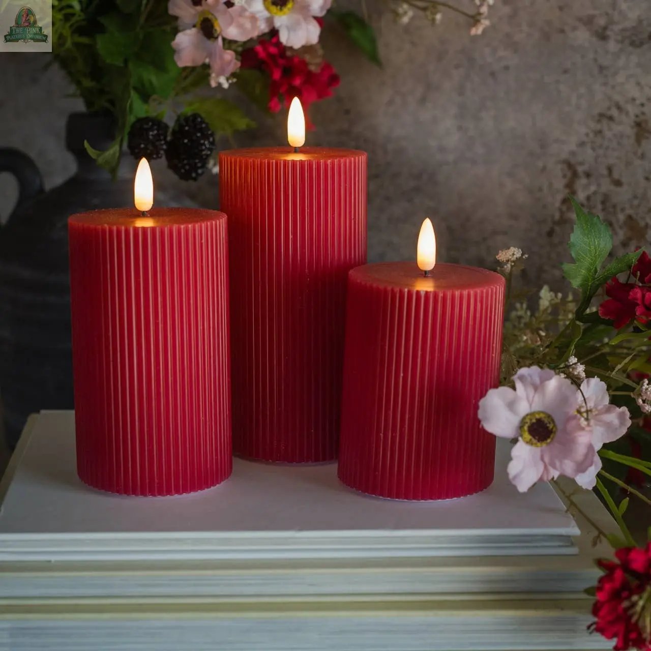 Three 6" RED GROOVED 3D FLAME CANDLES with glowing flames are arranged on stacked white books, surrounded by pink and red flowers and greenery—a charming Ragon House accent for any space.