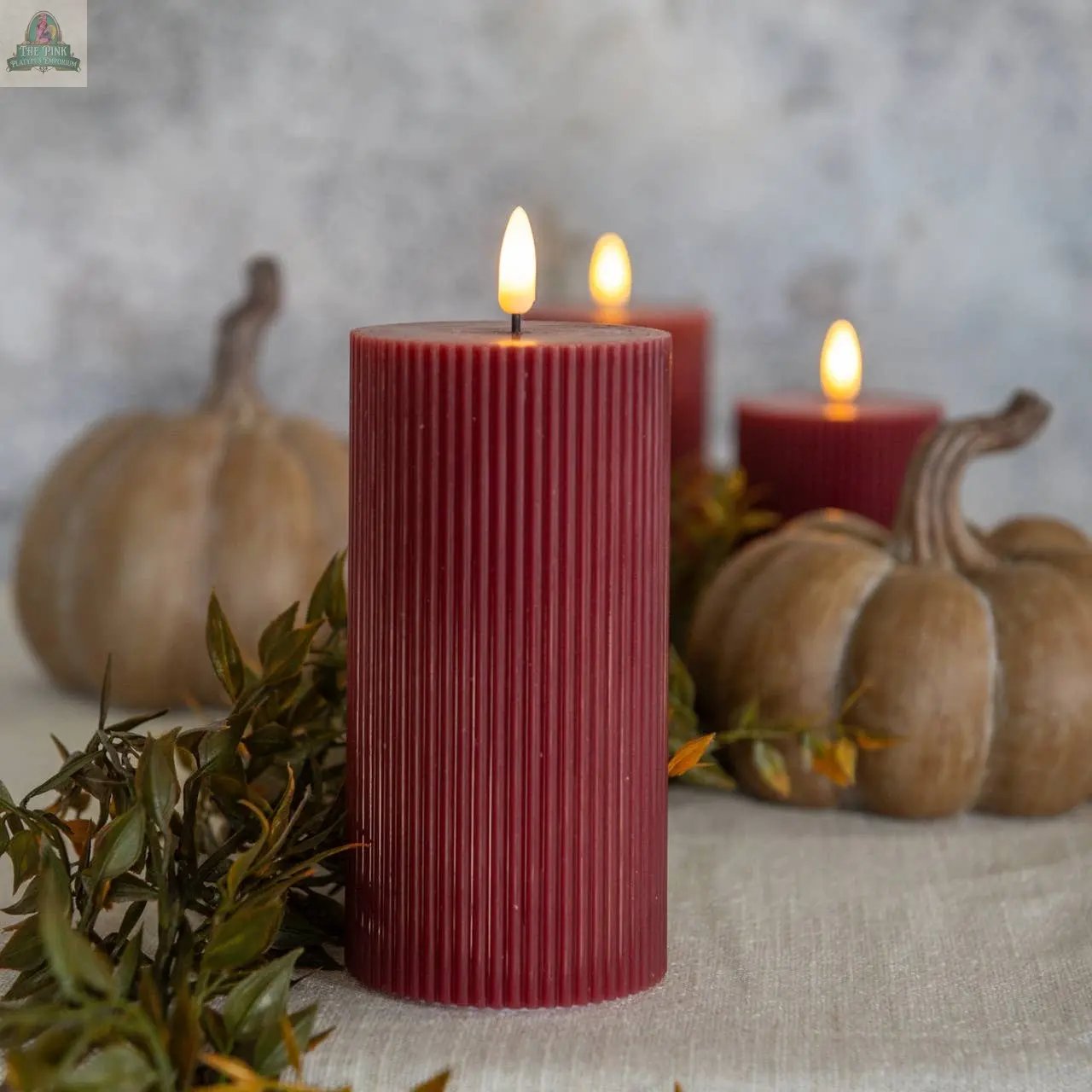 A 6" RED GROOVED 3D FLAME CANDLE burns beside two brown pumpkins and greenery, with two more red candles behind, all atop light fabric, creating a cozy, autumnal scene ideal for Ragon House décor.