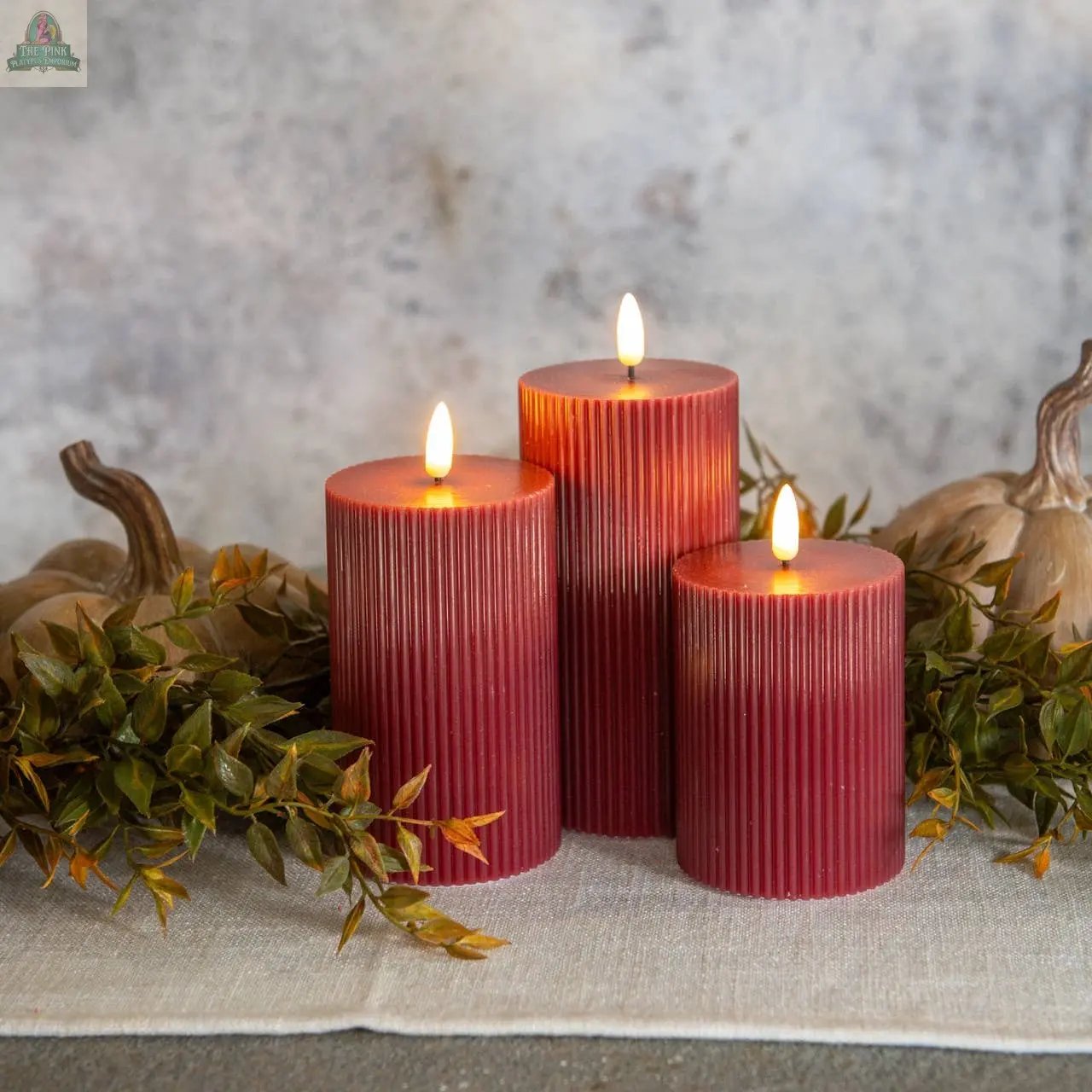 Three 6" RED GROOVED 3D FLAME CANDLES glow warmly on a beige cloth, surrounded by green leaves and brown gourds, set against a textured gray wall for a cozy autumn look.