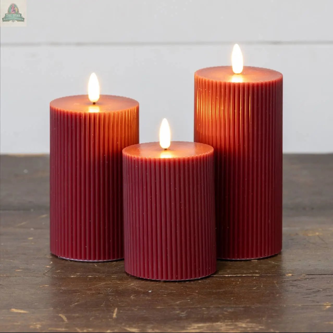 A trio of 6" RED GROOVED 3D FLAME CANDLES from Ragon House, lit on a wooden surface, showcases their elegant, ribbed design against a light backdrop.