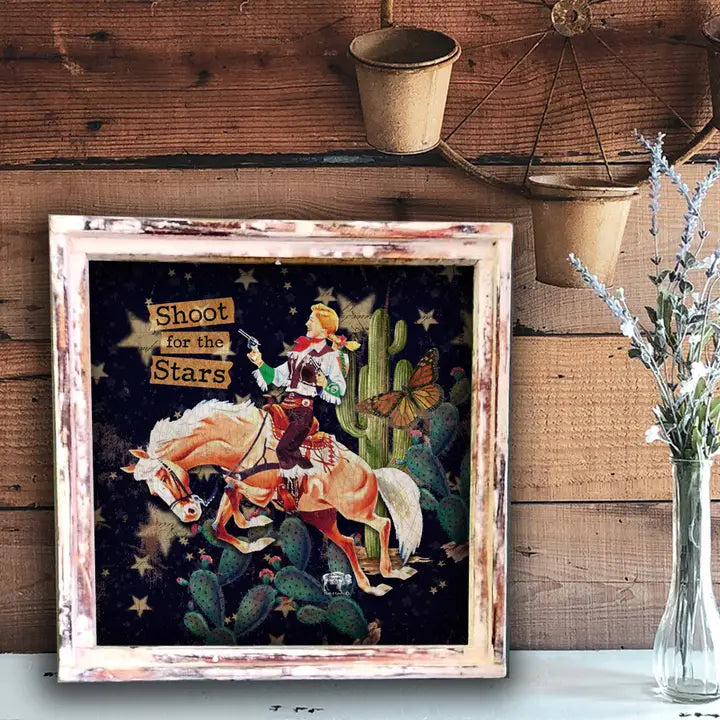 Framed artwork of a cowgirl on a horse with cacti and stars, placed on a wooden surface.