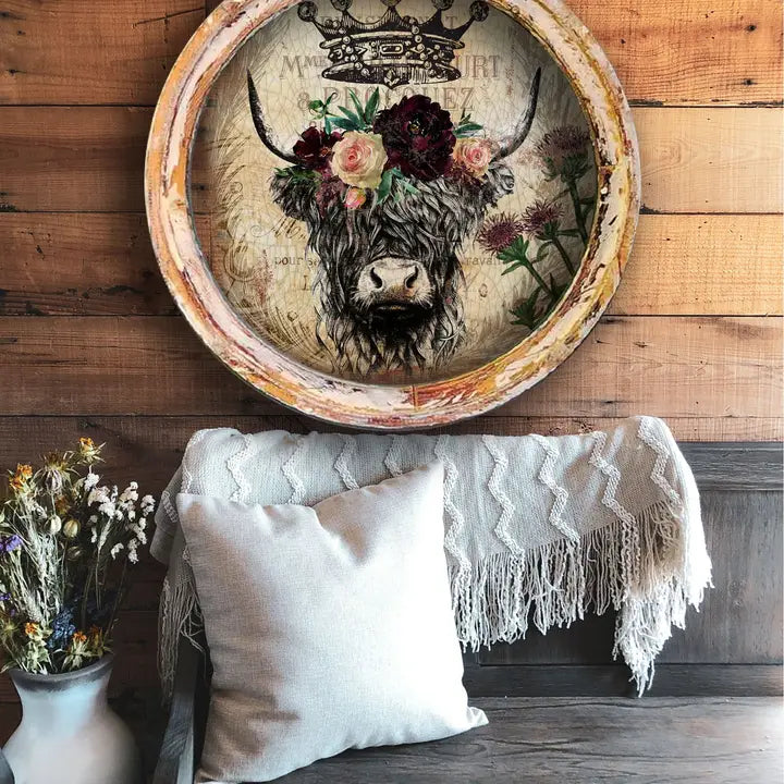 decorative farmhouse wall decor with a cow's head and floral crown on a wooden wall, below a white pillow and blanket.