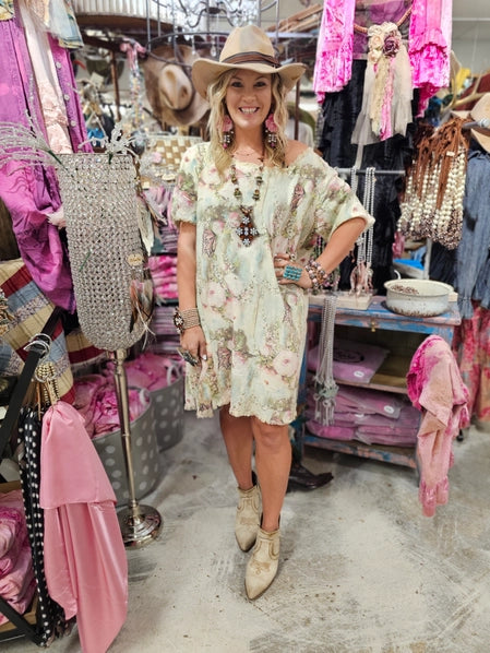 Woman in a boho western floral dress and cowboy hat standing in a clothing store with racks of clothes in the background.