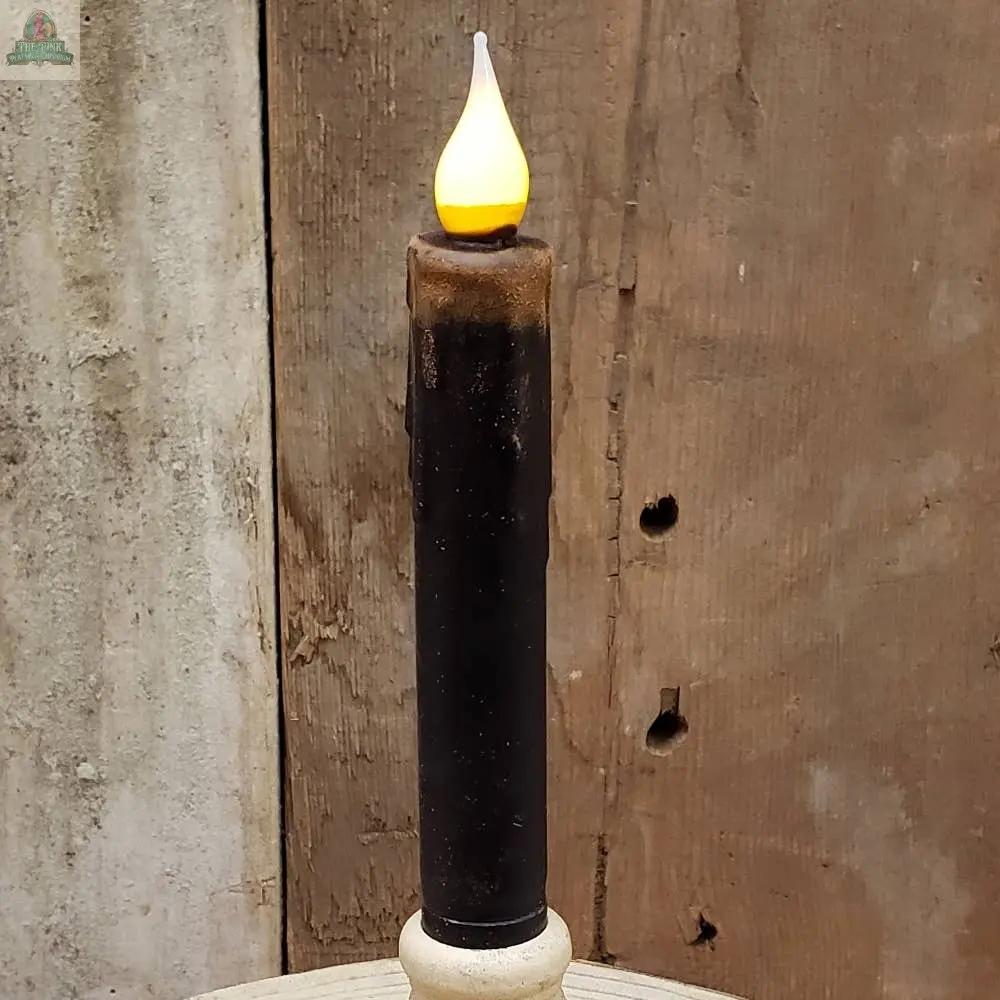 The 7" Timered Color Taper in Black, a battery-operated candle with a glowing flame, sits on a light surface against a weathered wooden background.