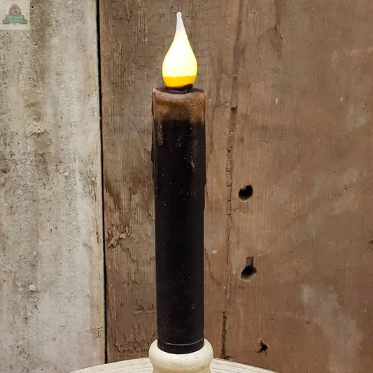The 7" Timered Color Taper in Black, a battery-operated candle with a glowing flame, sits on a light surface against a weathered wooden background.