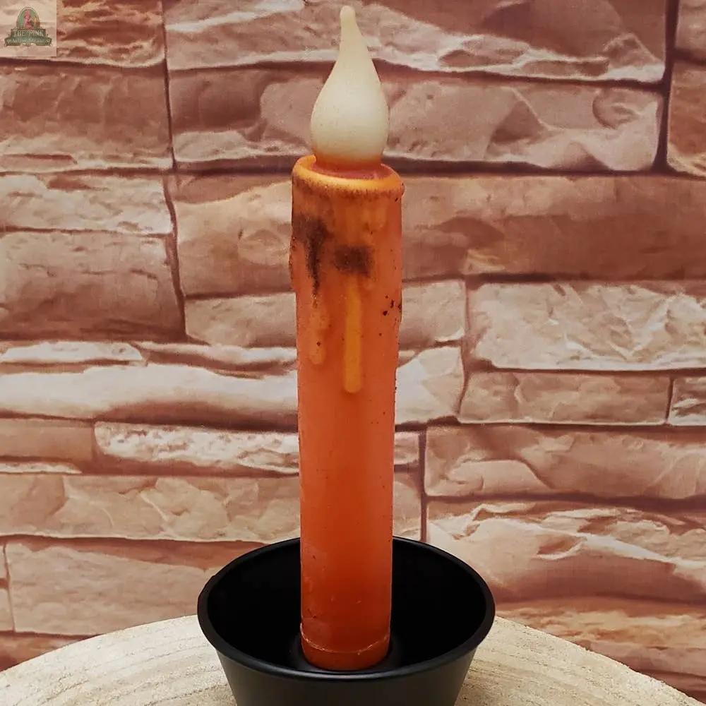 A 7" Timered Color Taper in black stands upright in a black holder, featuring a realistic flame-shaped top and wax drips, set against a stone wall background.