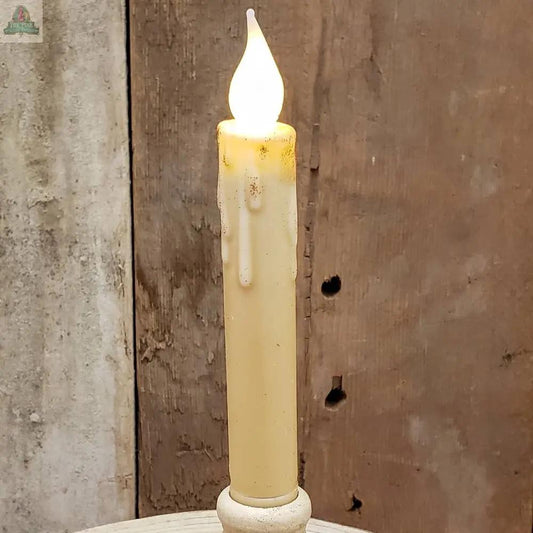 The 7" Timered Color Taper in cream stands upright with a glowing flame-shaped bulb, artificial wax drips, and a rustic wooden background.