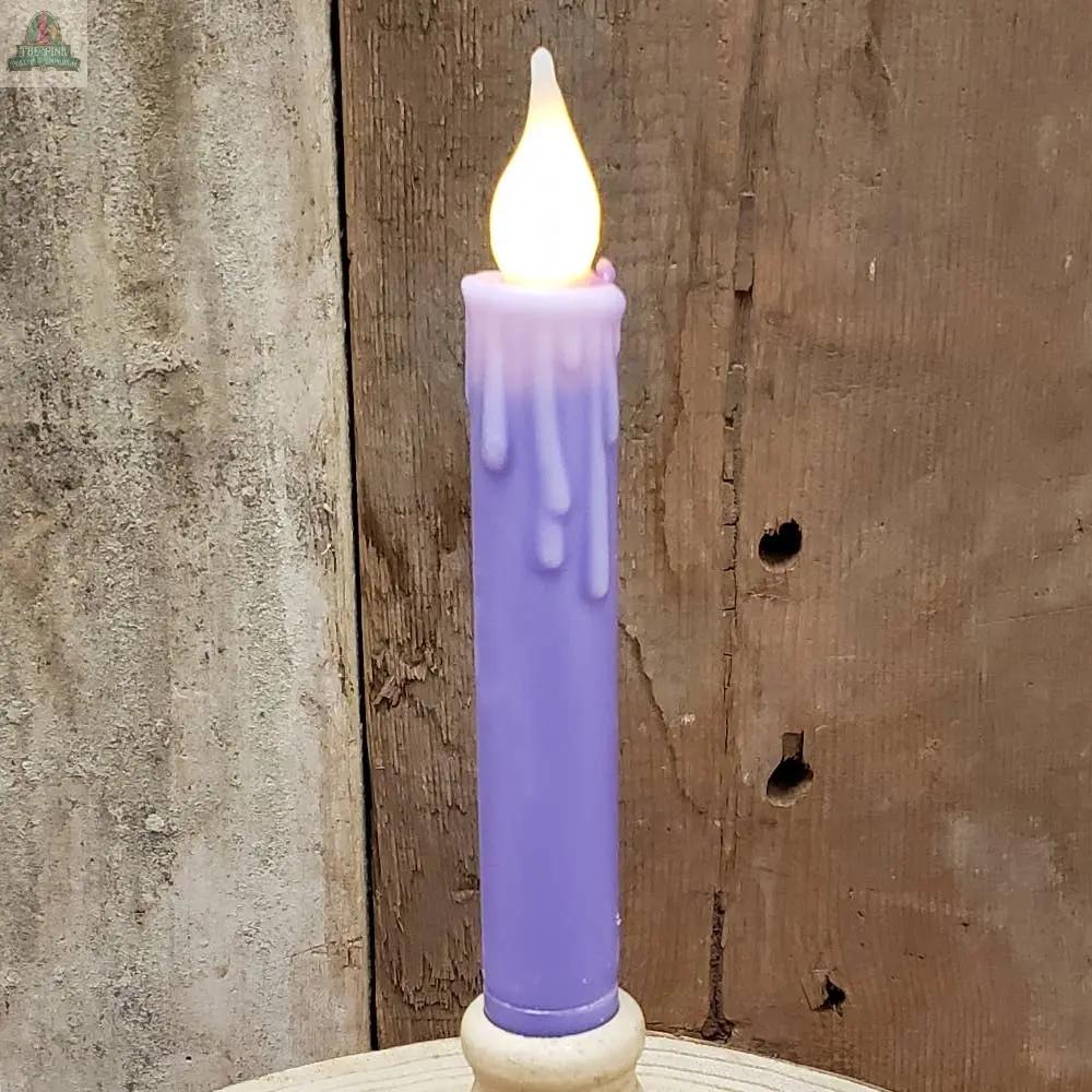 A tall, cream 7" Timered Color Taper candle with melting wax details and a glowing flame stands upright on a round wooden base in front of a rustic wooden wall.