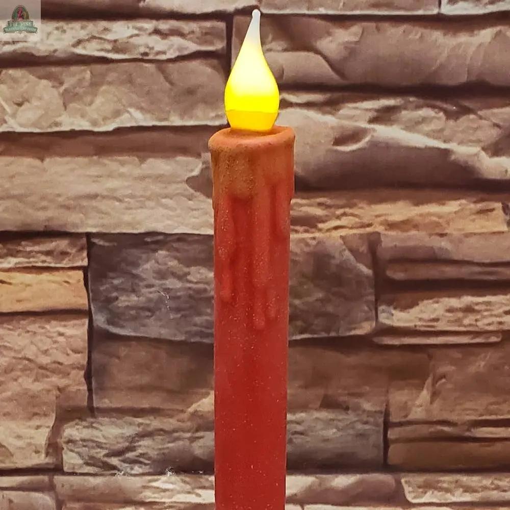 The 7" Timered Color Taper in red has a yellow flame-shaped LED and wax-like drips, creating a realistic effect as it sits before a textured stone wall.
