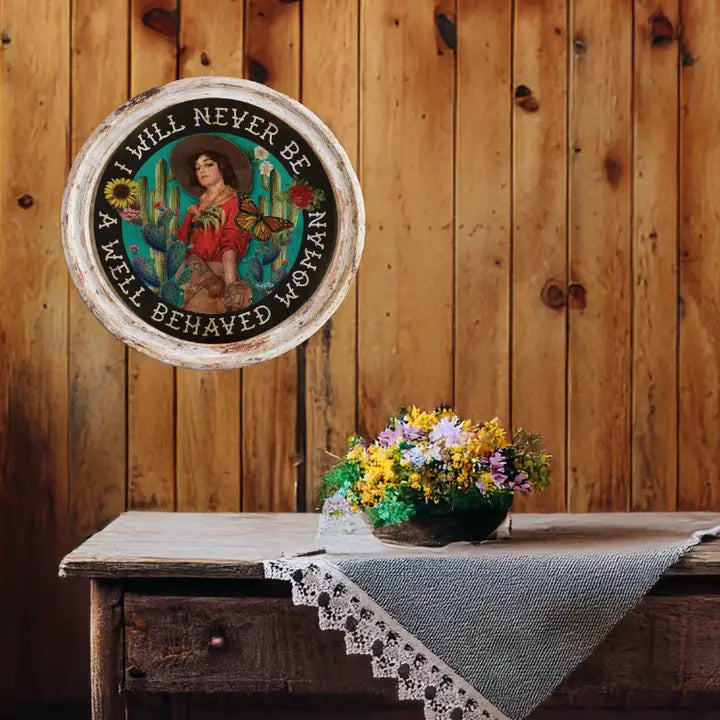 Western wall art with a woman and cacti on a wooden wall with a table and flowers in the foreground.