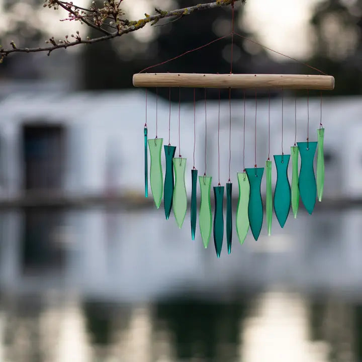 Decorative wind chime with green glass elements hanging from a branch over a blurred natural background.