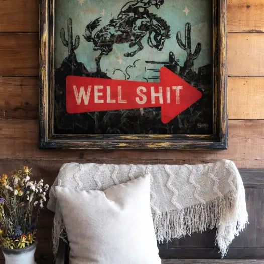 Framed artwork with a red arrow and text saying "well shit" with a cowboy on a horse on a wooden wall above a couch.