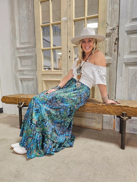 Woman sitting on a wooden bench wearing a white off-shoulder top Peacock Feather Boho Pants, with a hat.