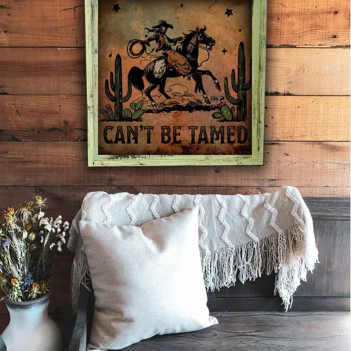 Framed artwork with a cowboy and horse graphic and 'Can't Be Tamed' text on a wooden wall above a couch.
