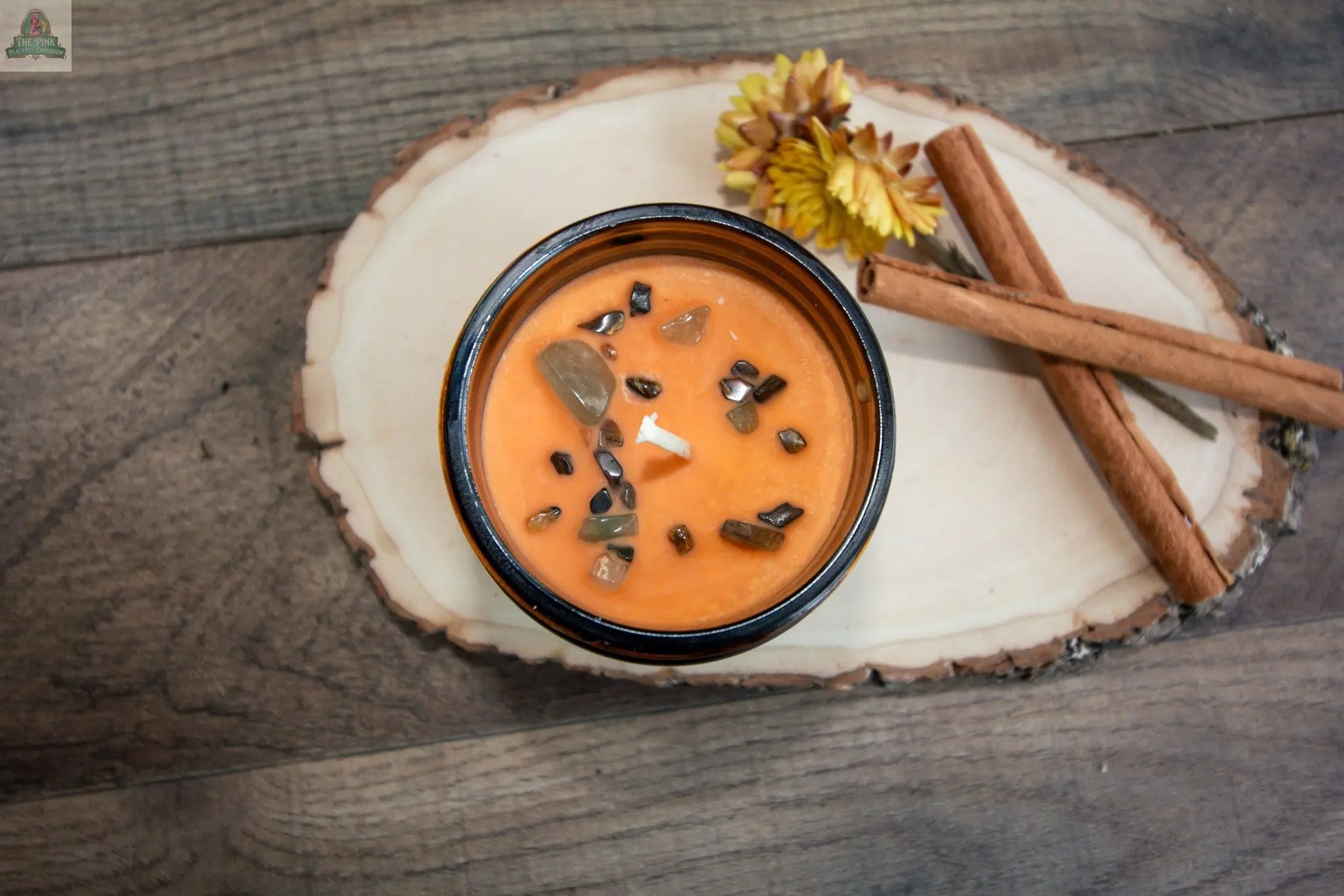 Abundance, a pumpkin soufflé candle with orange wax and decorative stones on top, rests on a wooden slice alongside two cinnamon sticks and dried yellow flowers, all arranged on a wooden surface.