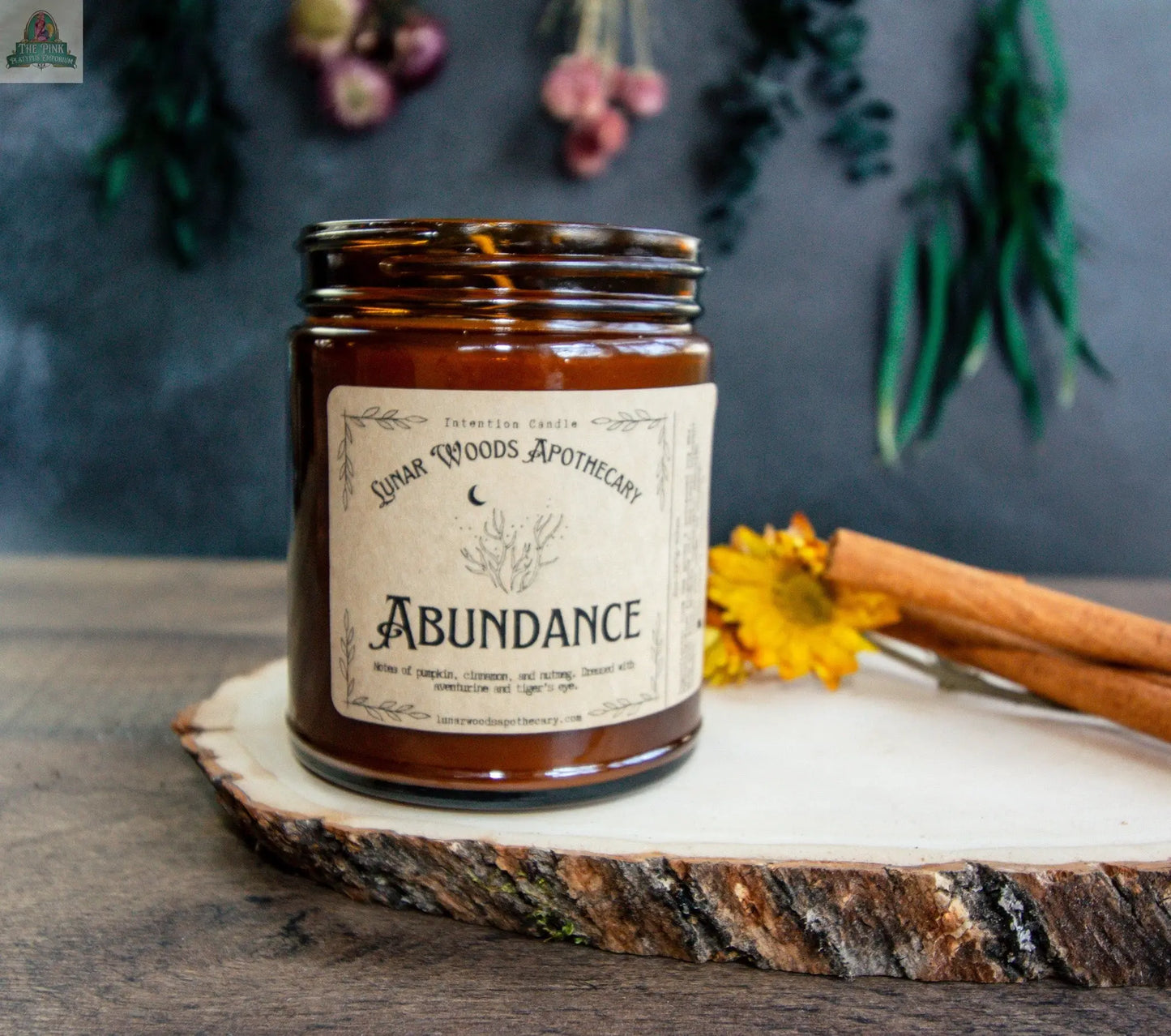 The Abundance candle, crafted from brown glass, rests on a wood slice with a yellow flower and cinnamon sticks nearby; dried flowers and greenery are softly blurred in the background.