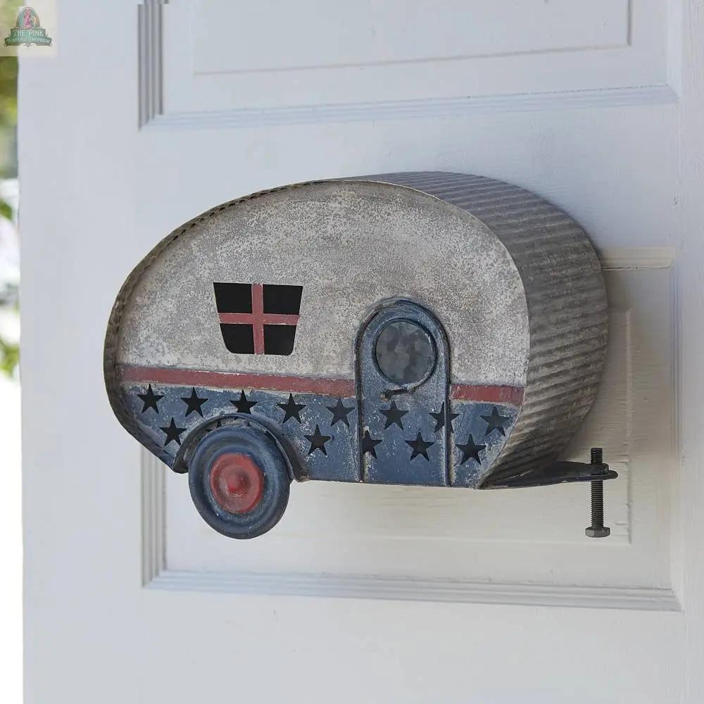 The Americana Camper Hanging Birdhouse is a metal birdhouse shaped like a retro camper, featuring star cutouts, a small door, a painted wheel, and a rustic finish, mounted on a white door.