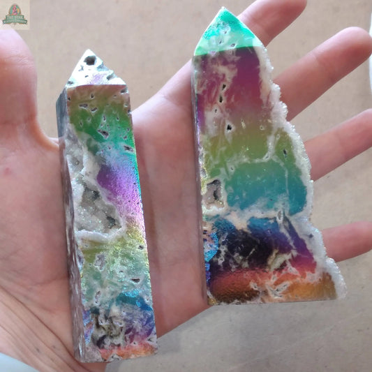 A hand holds two vibrant, marbled crystal towers from Keystone Crystals, including the Angel Aura Druzy Sphalerite Obelisk— a shimmering piece with rainbow hues, sparkling texture, and rough edges from Pink Platypus Emporium.