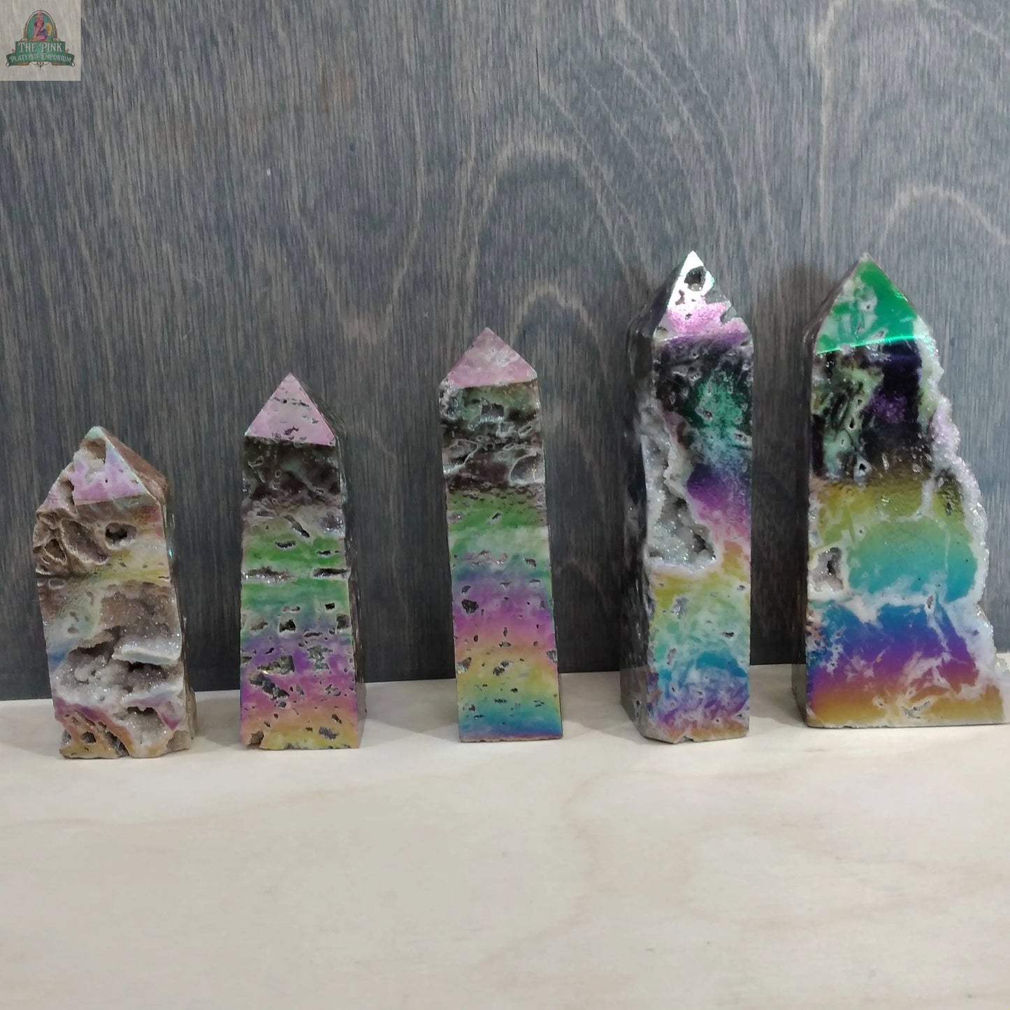 Five vibrant Angel Aura Druzy Sphalerite Obelisks from Keystone Crystals are displayed upright against a gray wooden backdrop. Each shimmering tower, available at Pink Platypus Emporium, features colorful hues and dazzling natural textures.
