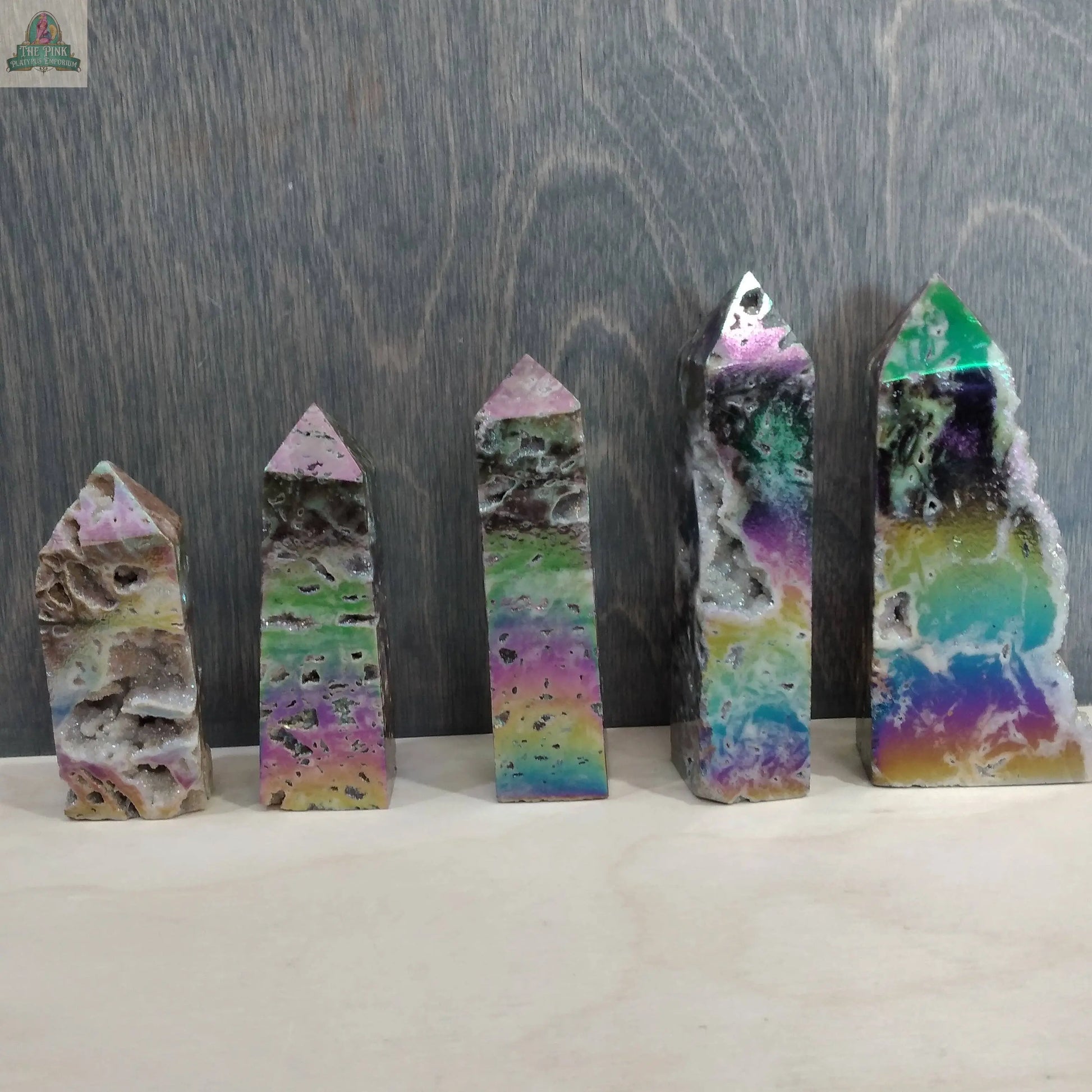 Five vibrant Angel Aura Druzy Sphalerite Obelisks from Keystone Crystals are displayed upright against a gray wooden backdrop. Each shimmering tower, available at Pink Platypus Emporium, features colorful hues and dazzling natural textures.