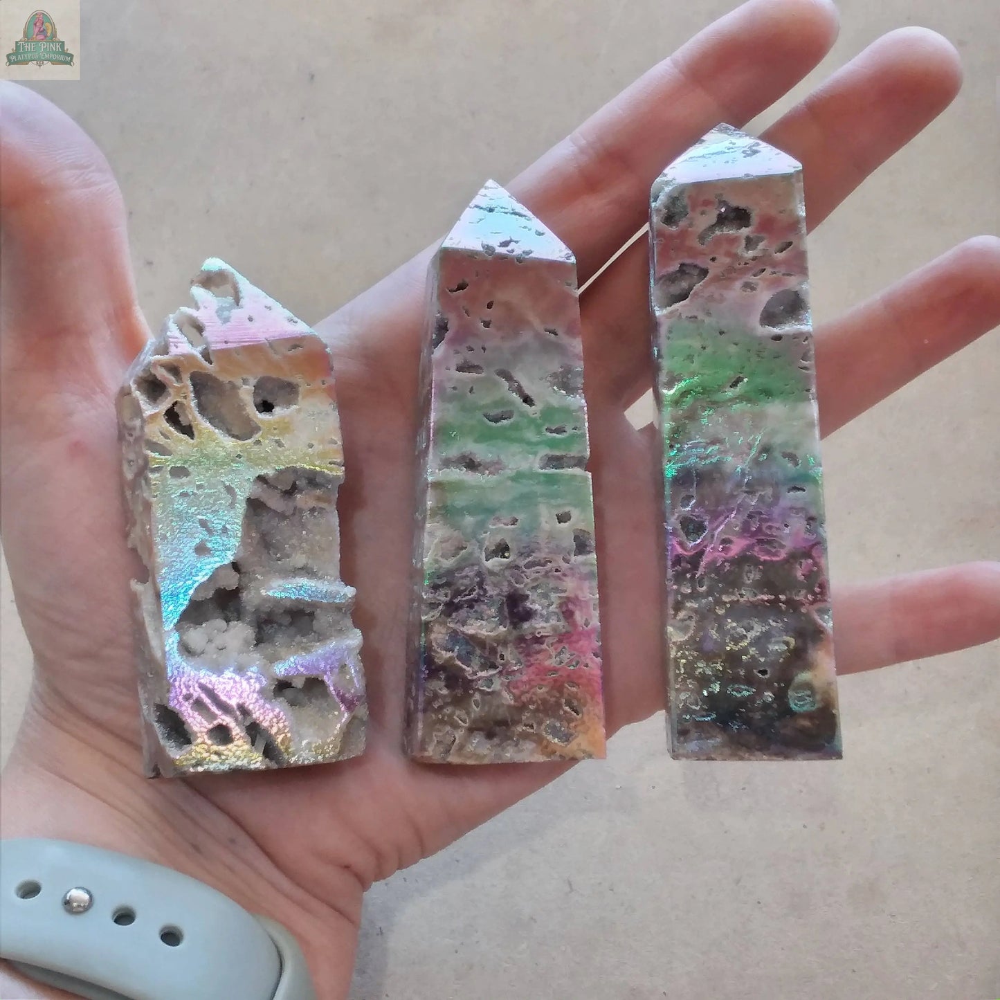 A hand holds three Angel Aura Druzy Sphalerite Obelisk – Shimmering Crystal Towers by Keystone Crystals, showing rough, multicolored surfaces in pink, green, and purple with a metallic sheen. Available at Pink Platypus Emporium.