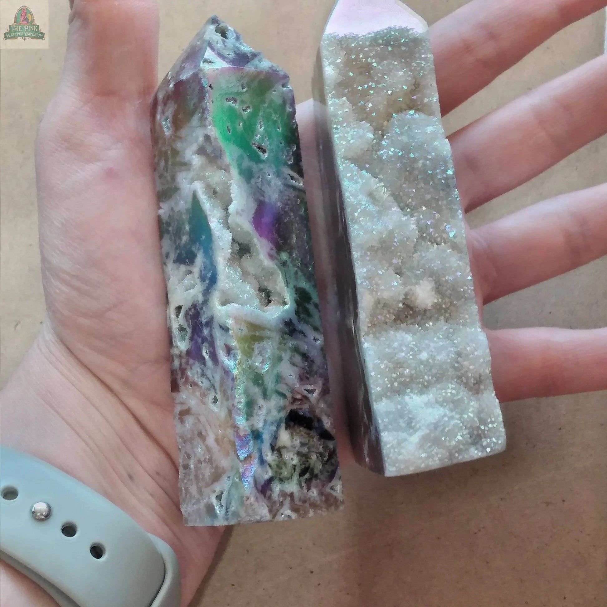 A hand holds two pointed crystal towers from Keystone Crystals: a vibrant green-and-purple Sphalerite obelisk and the Angel Aura Druzy Sphalerite Obelisk – Shimmering Crystal Tower, sparkling with druzy texture from Pink Platypus Emporium.