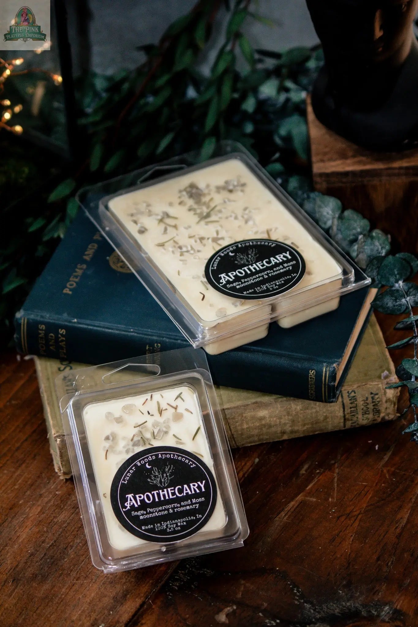 Two packages of Apothecary — Witchy Wax Melts in Lunar Woods scent with botanical ingredients rest on stacked vintage books, surrounded by greenery on a wooden surface.
