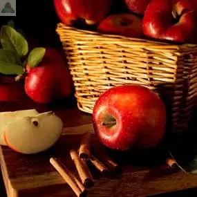 A bottle of Apple Cinnamon Atomizer Oils rests on a wooden surface, surrounded by fresh red apples, a sliced apple, cinnamon sticks, potpourri, and green leaves, creating a cozy, rustic atmosphere.