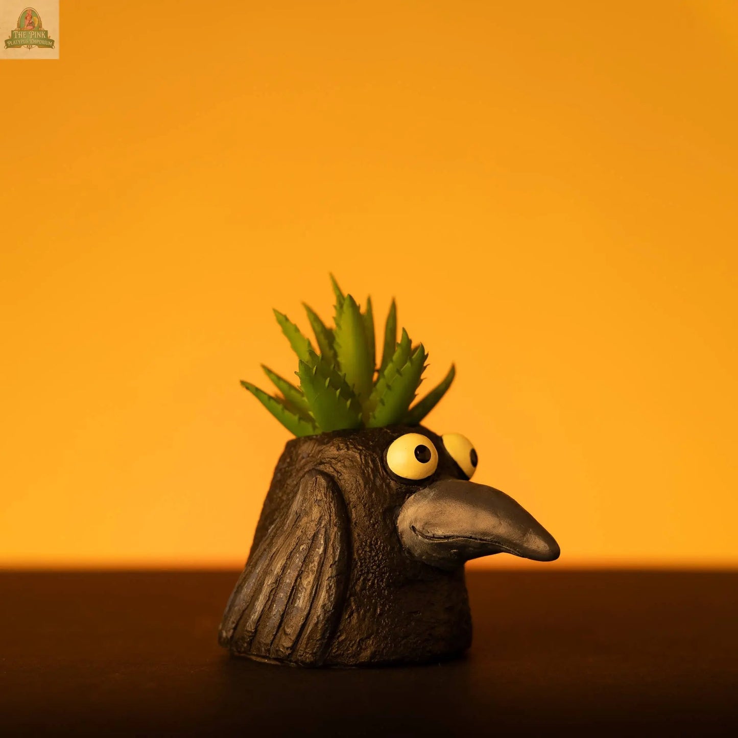The Baby Eddie Crow Blobhouse Planter, a small handcrafted pot shaped like a black bird’s head with big eyes and a long beak, holds a spiky green succulent—perfect for unique home décor against vibrant backgrounds.