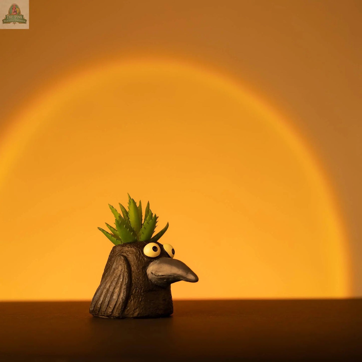 The Baby Eddie Crow Blobhouse Planter is a quirky poly-resin bird with a long beak, cartoonish eyes, and green succulent leaves on its head—a playful accent for home décor and small plants, glowing warmly on any surface.