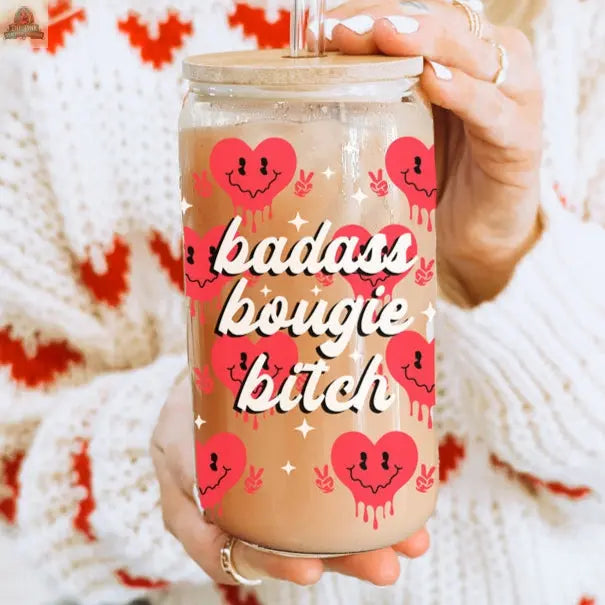 A person in a white and red sweater holds a Badass Bougie 20oz glass cup, filled with iced coffee in a mason jar decorated with red smiling drip-heart designs and bold white lettering.