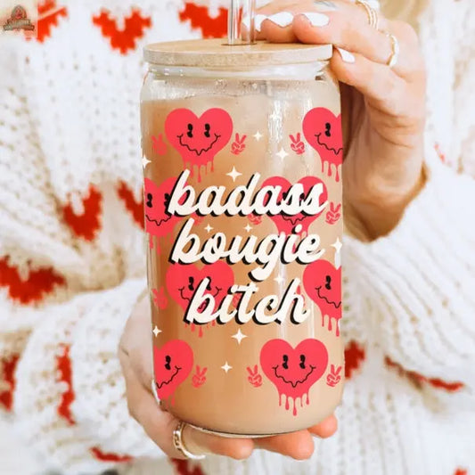 A person in a white and red sweater holds a Badass Bougie 20oz glass cup, filled with iced coffee in a mason jar decorated with red smiling drip-heart designs and bold white lettering.