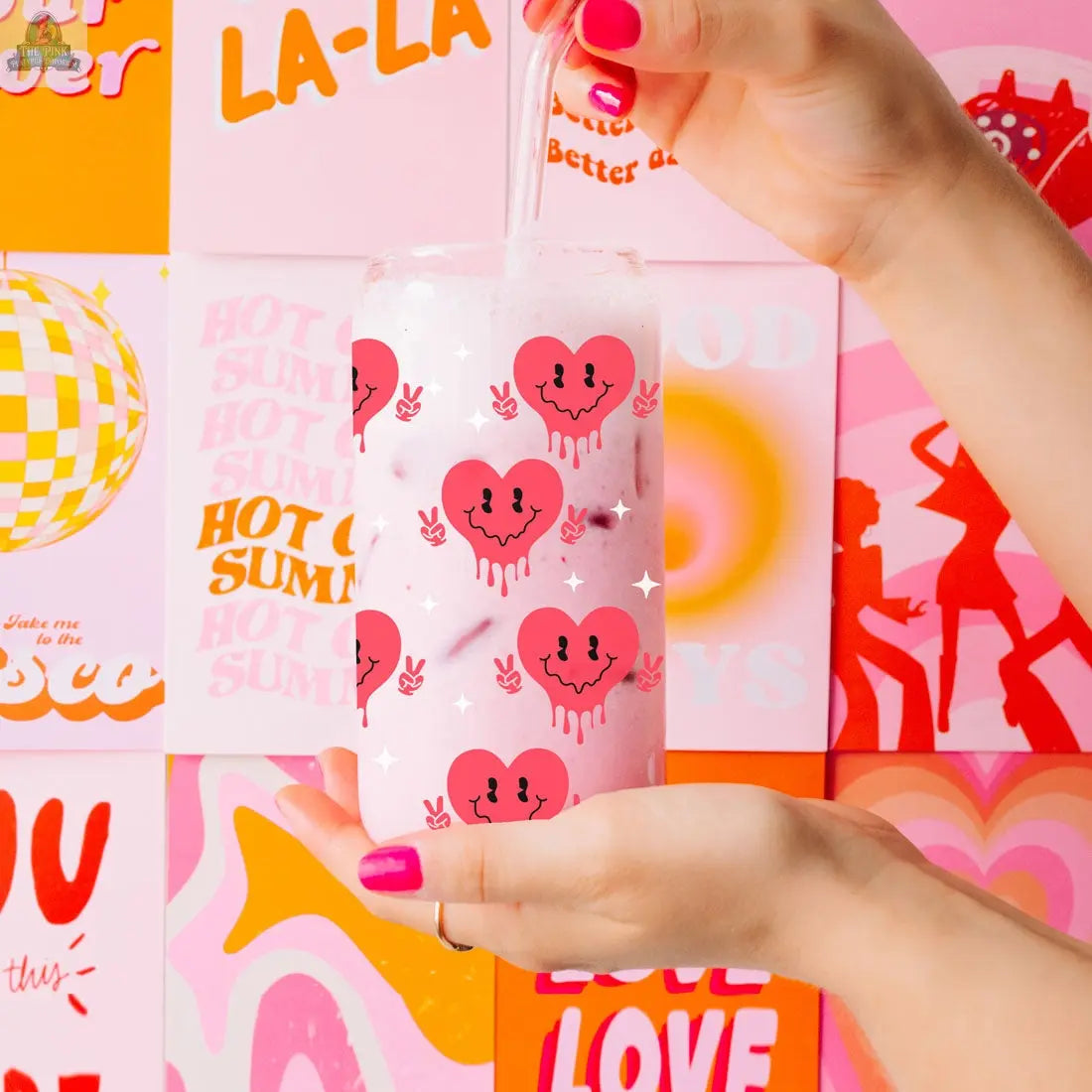 A hand with pink nail polish holds a Badass Bougie 20oz glass cup with a straw, featuring cute melting heart designs. Colorful retro-inspired posters with bold, vibrant graphics appear in the background.