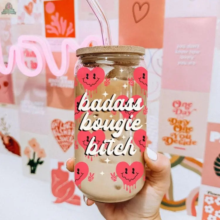 A hand holds the Badass Bougie 20oz glass cup filled with iced coffee, featuring a bamboo lid and straw. White script and pink hearts stand out against a blurred pink and orange art collage in the background.