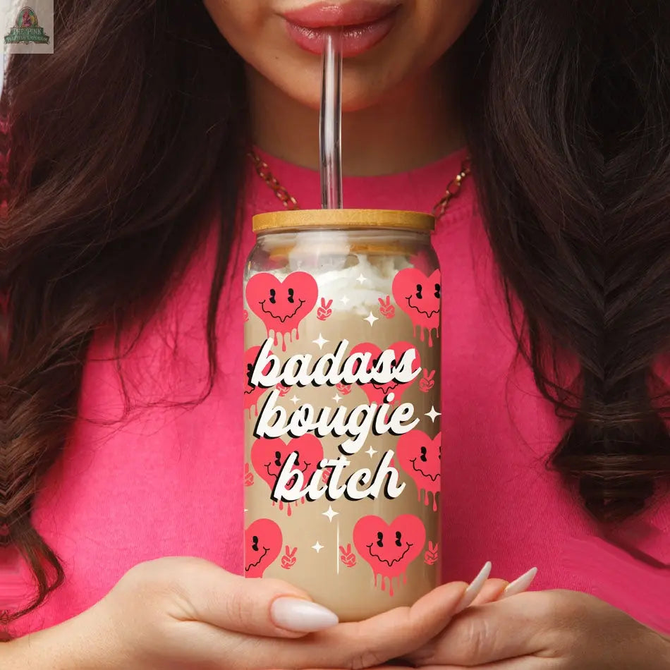 A woman in a pink top holds a Badass Bougie 20oz glass cup with a bamboo lid and metal straw, featuring pink hearts with smiling faces and playful dripping designs.