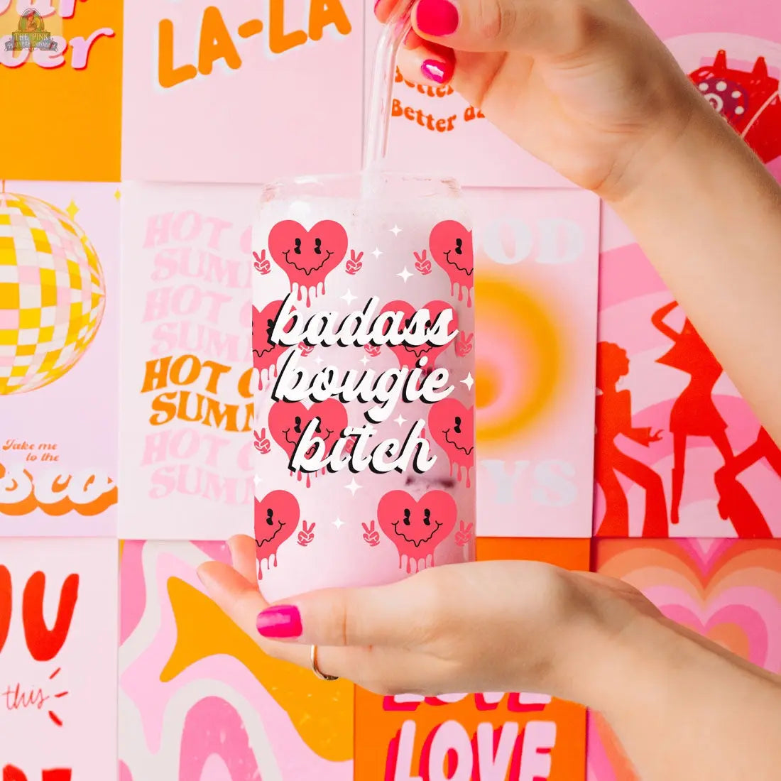 A hand with pink-painted nails holds a Badass Bougie 20oz glass cup with pink heart designs and white lettering, set against a colorful wall with retro-style posters and graphics.
