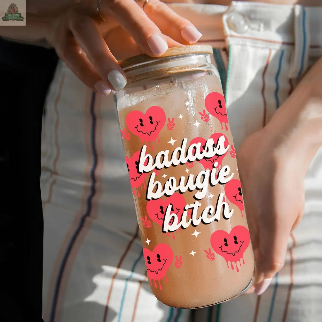 Someone holds a Badass Bougie 20oz glass cup with bold white lettering and pink smiling hearts. Their iced drink shines through the glass as they display their striped pants.