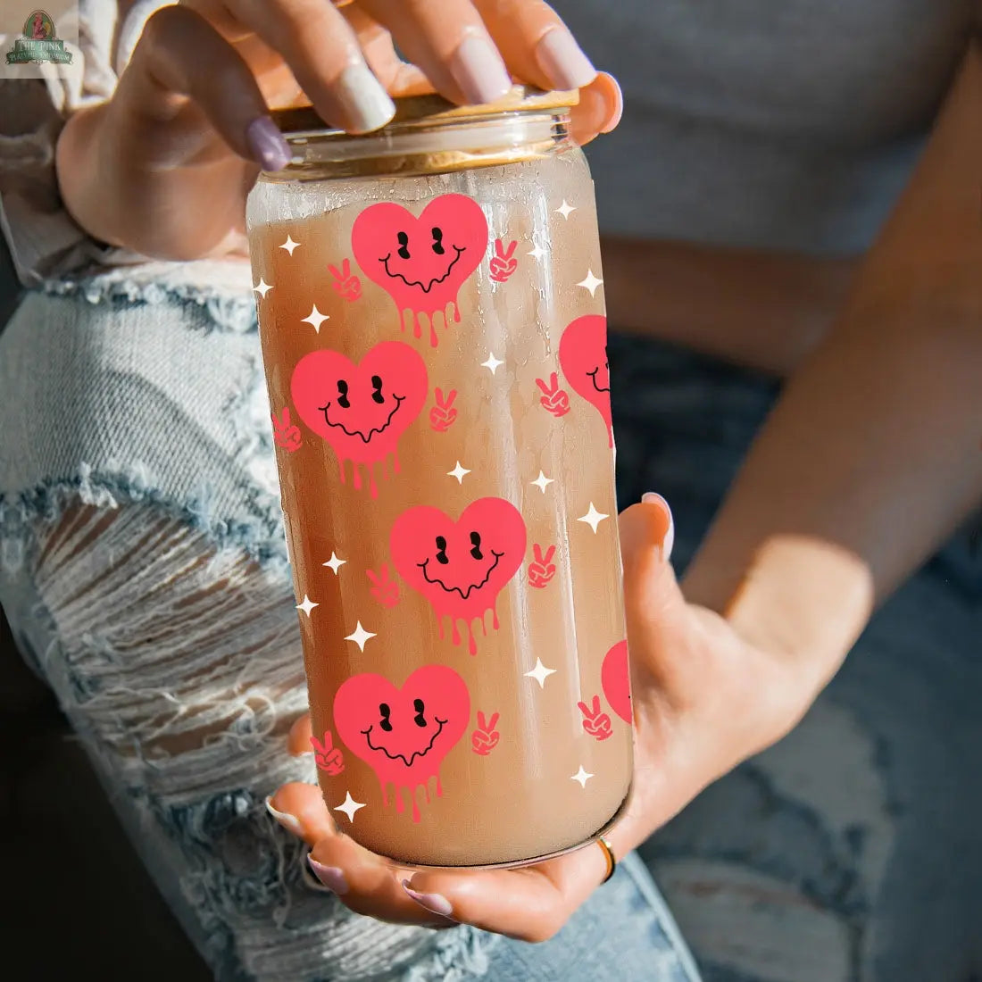 A person in ripped jeans holds a Badass Bougie 20oz glass cup with bamboo lid, featuring pink dripping heart faces, peace signs, and white star designs.