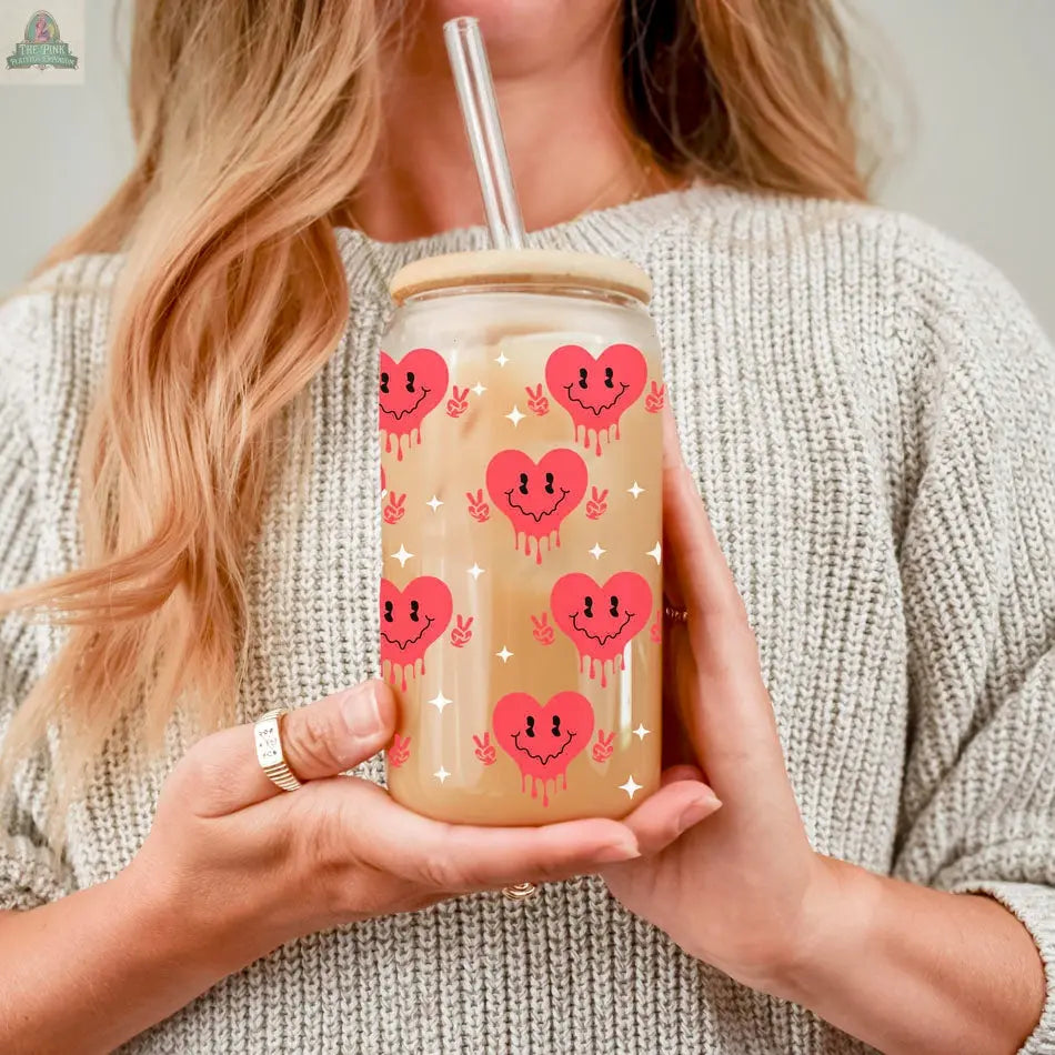 A woman with wavy blonde hair and rings, dressed in a light gray knitted sweater, holds the Badass Bougie 20oz glass cup with a bamboo lid and clear straw, featuring red smiling heart faces and stars.