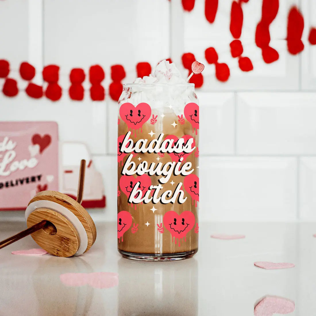 A Badass Bougie 20oz glass cup filled with iced coffee and whipped cream sits on a counter, adorned with pink smiling hearts. Red heart garland and heart-shaped decor add playful, festive accents to the scene.